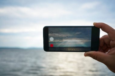 Gökyüzünde ve deniz geçmişinde akıllı bir telefonu tutan bir adam. Bulutların arasından ışık