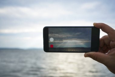 Gökyüzünde ve deniz geçmişinde akıllı bir telefonu tutan bir adam. Bulutların arasından ışık