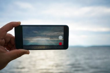 Gökyüzünde ve deniz geçmişinde akıllı bir telefonu tutan bir adam. Bulutların arasından ışık