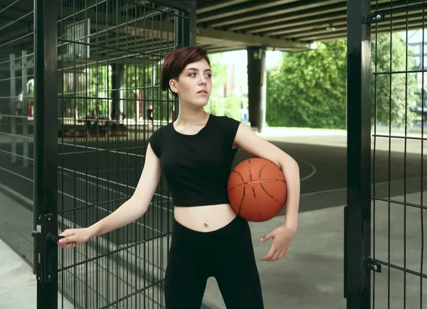 caucasian girl standing near fence of outdoor street basketball court, holding a ball. short haircut