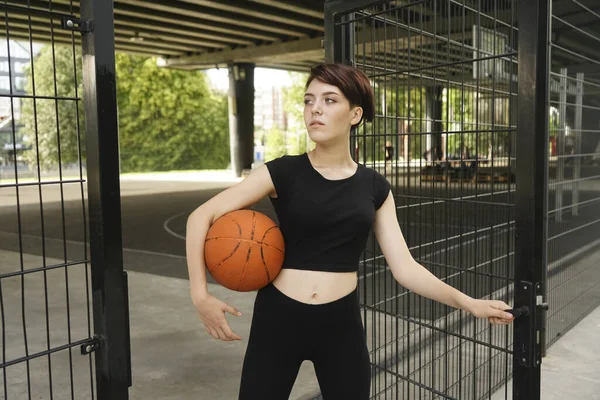 caucasian girl standing near fence of outdoor street basketball court, holding a ball. short haircut