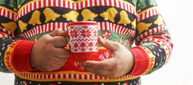 Sıcak çikolata içen bir adam. Noel ağacı deseni. Madalyalı içki kupası. Erkek, kırmızı fincanda sıcak kahve tutan örgü süveter ve Xmas süsü giyiyor. Eşcinseller kış tatillerini kutlar. Yüzü yok