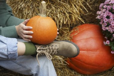 Kadın saman zeminde kot pantolonun içinde oturuyor ve ayakkabıları arasında balkabağı var. Portakal kabağı ayakkabının yanında. Sonbahar hasat konsepti. Komik sonbahar konsepti. resmi kapat