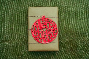 Kahverengi el sanatları kağıdına sarılı hediye kutusu. Yazılı kırmızı tahta oyuncak figürü - Mutlu Noeller. kırmızı ve beyaz ip. Çuval bezi arkaplanı.