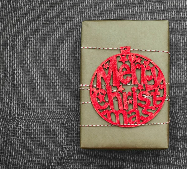 礼品盒用褐色手工纸包好 红色木制玩具人像 上面有题词 快乐的圣诞节 红色和白色的绳子 麻布背景 — 图库照片