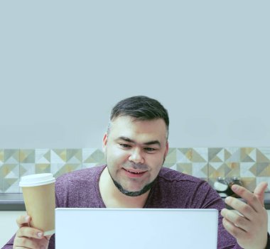 Orta yaşlı adam evde çalışıyor ve dizüstü bilgisayar kullanıyor. Erkek kahve içiyor ve bilgisayar ekranına bakıyor. Gülen surat. Arka planda mutfak içi.