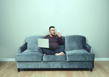 Orta yaşlı bir adam evdeki koltukta otururken dizüstü bilgisayar ve cep telefonu kullanıyor. meşgul Asyalı sakallı adam gri koltukta oturuyor ve cep telefonu ve dizüstü bilgisayarla çalışıyor..