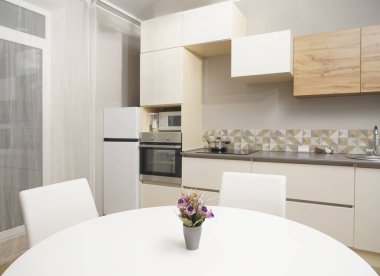Mutfağa yakın vazolu yemek masası buzdolabı, zemin tasarım, elektrik, soba duvarda asılı vazo, yemek masasında çiçek saksısı ve seramik tabak, beyaz renkli sandalye.