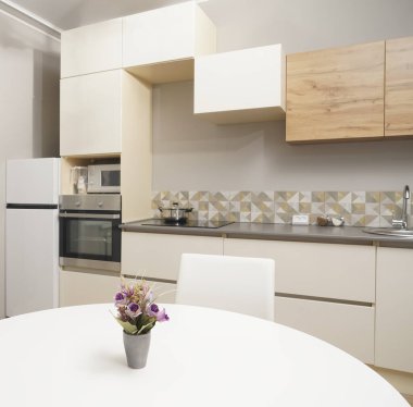 Mutfağa yakın vazolu yemek masası buzdolabı, zemin tasarım, elektrik, soba duvarda asılı vazo, yemek masasında çiçek saksısı ve seramik tabak, beyaz renkli sandalye.