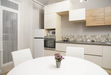 Mutfağa yakın vazolu yemek masası buzdolabı, zemin tasarım, elektrik, soba duvarda asılı vazo, yemek masasında çiçek saksısı ve seramik tabak, beyaz renkli sandalye.