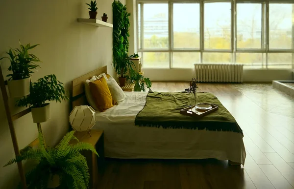 King size bed with white and green bedding, large window and a painting on the wall in a bright hotel room interior. green plants around bed.