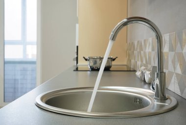 Modern tasarımlı krom musluk paslanmaz çelik mutfak lavabosunun üzerinde. Parlak beyaz mutfağın içi. Su açık bir musluktan dökülüyor.