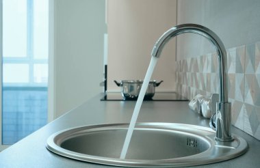 Modern tasarımlı krom musluk paslanmaz çelik mutfak lavabosunun üzerinde. Parlak beyaz mutfağın içi. Su açık bir musluktan dökülüyor.