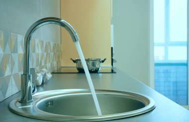 Modern tasarımlı krom musluk paslanmaz çelik mutfak lavabosunun üzerinde. Parlak beyaz mutfağın içi. Su açık bir musluktan dökülüyor.
