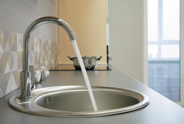 Modern tasarımlı krom musluk paslanmaz çelik mutfak lavabosunun üzerinde. Parlak beyaz mutfağın içi. Su açık bir musluktan dökülüyor.