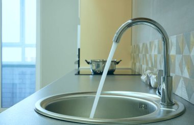 Modern tasarımlı krom musluk paslanmaz çelik mutfak lavabosunun üzerinde. Parlak beyaz mutfağın içi. Su açık bir musluktan dökülüyor.