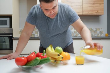 Orta yaşlı bir adam masada taze sıkılmış meyve suyu yapıyor. meyve ve sebze. Bir şişe ve bir bardak portakal suyu. Erkek, şişeden cama portakal suyu döküyor..