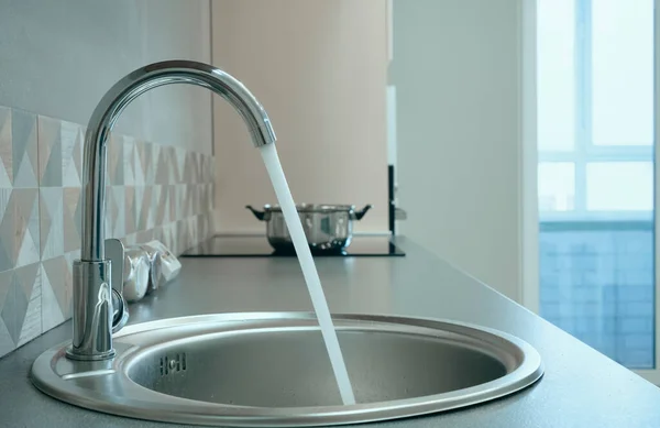 Modern designer chrome water tap over stainless steel kitchen sink. Interior of bright white kitchen. Water is poured through an open tap