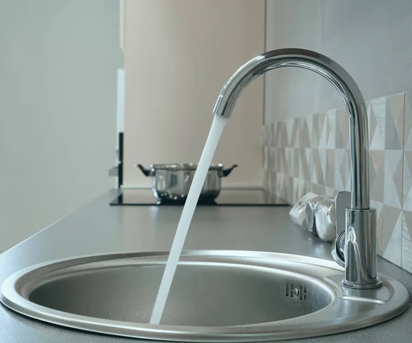 Modern designer chrome water tap over stainless steel kitchen sink. Interior of bright white kitchen. Water is poured through an open tap