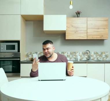 Orta yaşlı adam evde çalışıyor ve cep telefonu ve dizüstü bilgisayar kullanıyor. Erkek kahve içiyor ve bilgisayar ekranına bakıyor. Gülen surat. Arka planda mutfak içi.