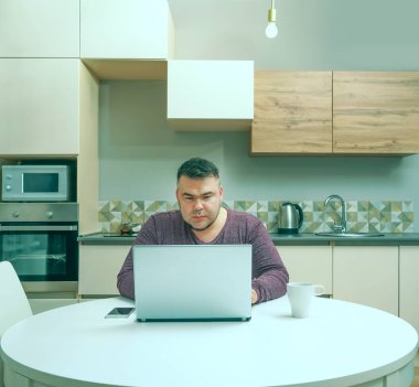 Orta yaşlı adam evde çalışıyor ve cep telefonu ve dizüstü bilgisayar kullanıyor. Erkek bilgisayar ekranına bakıyor. Ciddi bir yüz. Arka planda mutfak içi.