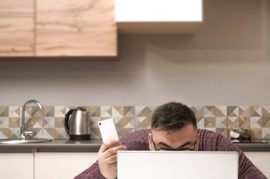 Orta yaşlı adam evde çalışıyor ve cep telefonu ve dizüstü bilgisayar kullanıyor. Erkek bilgisayar ekranına bakıyor. Ciddi bir yüz. Arka planda mutfak içi.