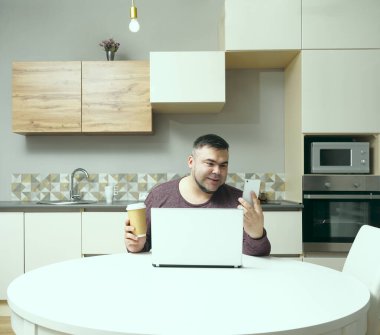Orta yaşlı adam evde çalışıyor ve cep telefonu ve dizüstü bilgisayar kullanıyor. Erkek kahve içiyor ve bilgisayar ekranına bakıyor. Gülen surat. Arka planda mutfak içi.