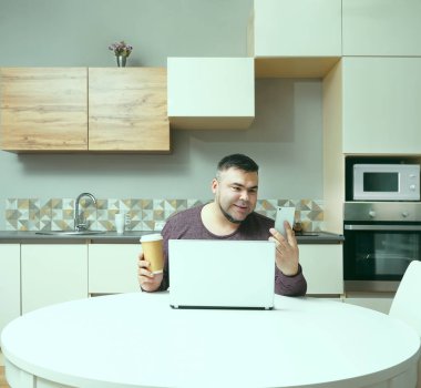 Orta yaşlı adam evde çalışıyor ve cep telefonu ve dizüstü bilgisayar kullanıyor. Erkek kahve içiyor ve bilgisayar ekranına bakıyor. Gülen surat. Arka planda mutfak içi.