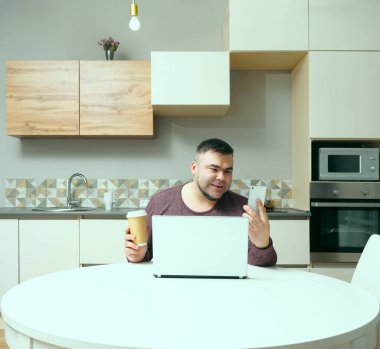 Orta yaşlı adam evde çalışıyor ve cep telefonu ve dizüstü bilgisayar kullanıyor. Erkek kahve içiyor ve bilgisayar ekranına bakıyor. Gülen surat. Arka planda mutfak içi.