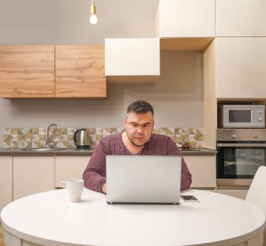 Orta yaşlı adam evde çalışıyor ve cep telefonu ve dizüstü bilgisayar kullanıyor. Erkek bilgisayar ekranına bakıyor. Ciddi bir yüz. Arka planda mutfak içi.