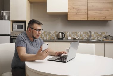 Kafkasyalı bir adam mutfakta cep telefonuyla sohbet ediyor ve dizüstü bilgisayarda çalışıyor. Modern mutfak iç mimarisi.