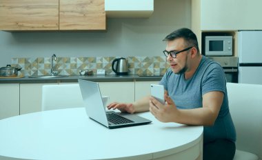 Kafkasyalı bir adam mutfakta cep telefonuyla sohbet ediyor ve dizüstü bilgisayarda çalışıyor. Modern mutfak iç mimarisi.