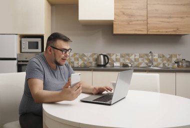 Kafkasyalı bir adam mutfakta cep telefonuyla sohbet ediyor ve dizüstü bilgisayarda çalışıyor. Modern mutfak iç mimarisi.