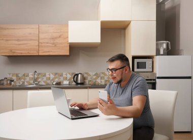 Kafkasyalı bir adam mutfakta cep telefonuyla sohbet ediyor ve dizüstü bilgisayarda çalışıyor. Modern mutfak iç mimarisi.