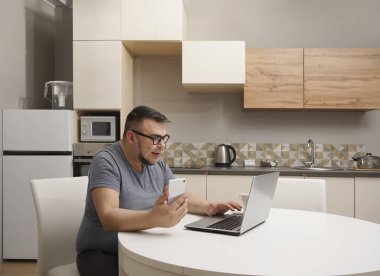 Kafkasyalı bir adam mutfakta cep telefonuyla sohbet ediyor ve dizüstü bilgisayarda çalışıyor. Modern mutfak iç mimarisi.