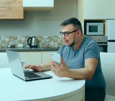 Kafkasyalı bir adam mutfakta cep telefonuyla sohbet ediyor ve dizüstü bilgisayarda çalışıyor. Modern mutfak iç mimarisi.