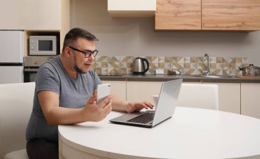 Kafkasyalı bir adam mutfakta cep telefonuyla sohbet ediyor ve dizüstü bilgisayarda çalışıyor. Modern mutfak iç mimarisi.