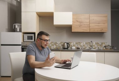 Kafkasyalı bir adam mutfakta cep telefonuyla sohbet ediyor ve dizüstü bilgisayarda çalışıyor. Modern mutfak iç mimarisi.