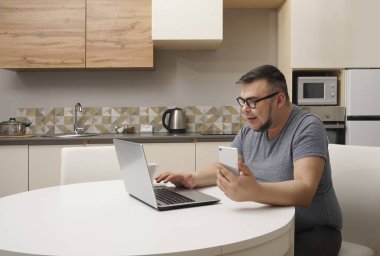 Kafkasyalı bir adam mutfakta cep telefonuyla sohbet ediyor ve dizüstü bilgisayarda çalışıyor. Modern mutfak iç mimarisi.