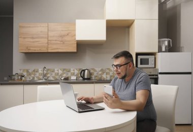 Kafkasyalı bir adam mutfakta cep telefonuyla sohbet ediyor ve dizüstü bilgisayarda çalışıyor. Modern mutfak iç mimarisi.