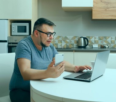 Kafkasyalı bir adam mutfakta cep telefonuyla sohbet ediyor ve dizüstü bilgisayarda çalışıyor. Modern mutfak iç mimarisi.