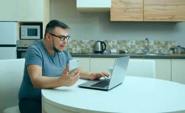 Kafkasyalı bir adam mutfakta cep telefonuyla sohbet ediyor ve dizüstü bilgisayarda çalışıyor. Modern mutfak iç mimarisi.