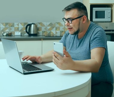 Kafkasyalı bir adam mutfakta cep telefonuyla sohbet ediyor ve dizüstü bilgisayarda çalışıyor. Modern mutfak iç mimarisi.