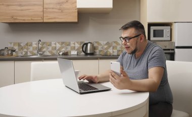 Kafkasyalı bir adam mutfakta cep telefonuyla sohbet ediyor ve dizüstü bilgisayarda çalışıyor. Modern mutfak iç mimarisi.