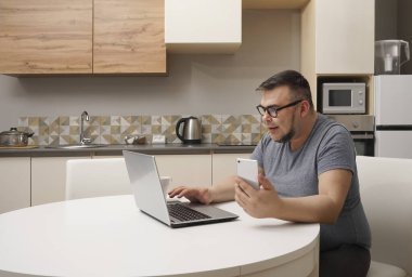 Kafkasyalı bir adam mutfakta cep telefonuyla sohbet ediyor ve dizüstü bilgisayarda çalışıyor. Modern mutfak iç mimarisi.