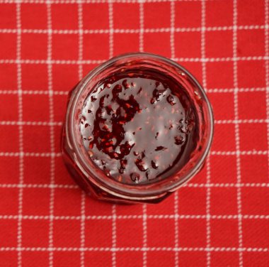 Kırmızı masa örtüsünde izole edilmiş kavanozdaki ahududu reçeli.