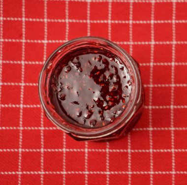 Kırmızı masa örtüsünde izole edilmiş kavanozdaki ahududu reçeli.