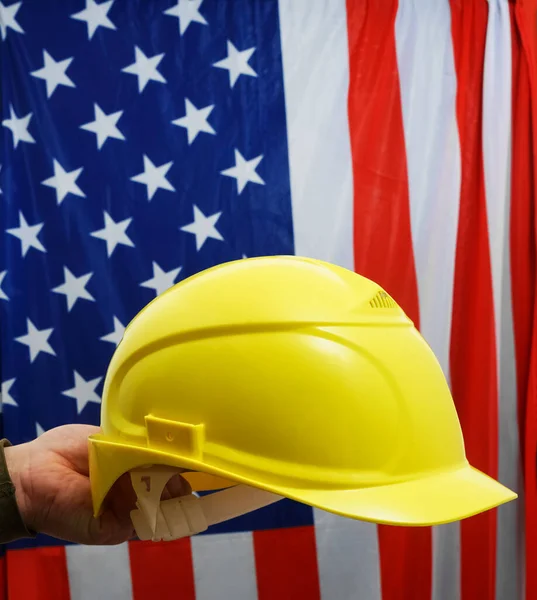 Hand  of engineer hold yellow plastic helmet for worker. American flag on background. happy  Labor day concept