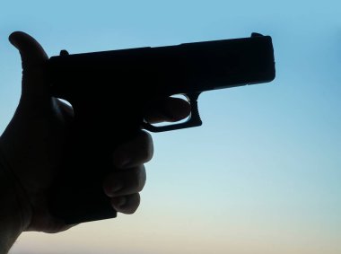 Mavi gökyüzü arka planında erkek eli silah tutuyor. Siyah tabanca silueti. 