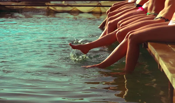 Group of women sitting in a row at the pool side. no faces. Group of girls sitting on a wooden platform by the river and splashing their feet in water on warm summer day. summertime season. beach party.